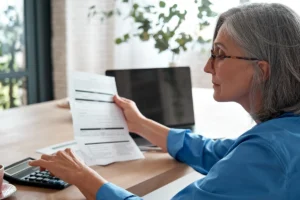 Mature woman holding paper bill using calculator managing finances at home.